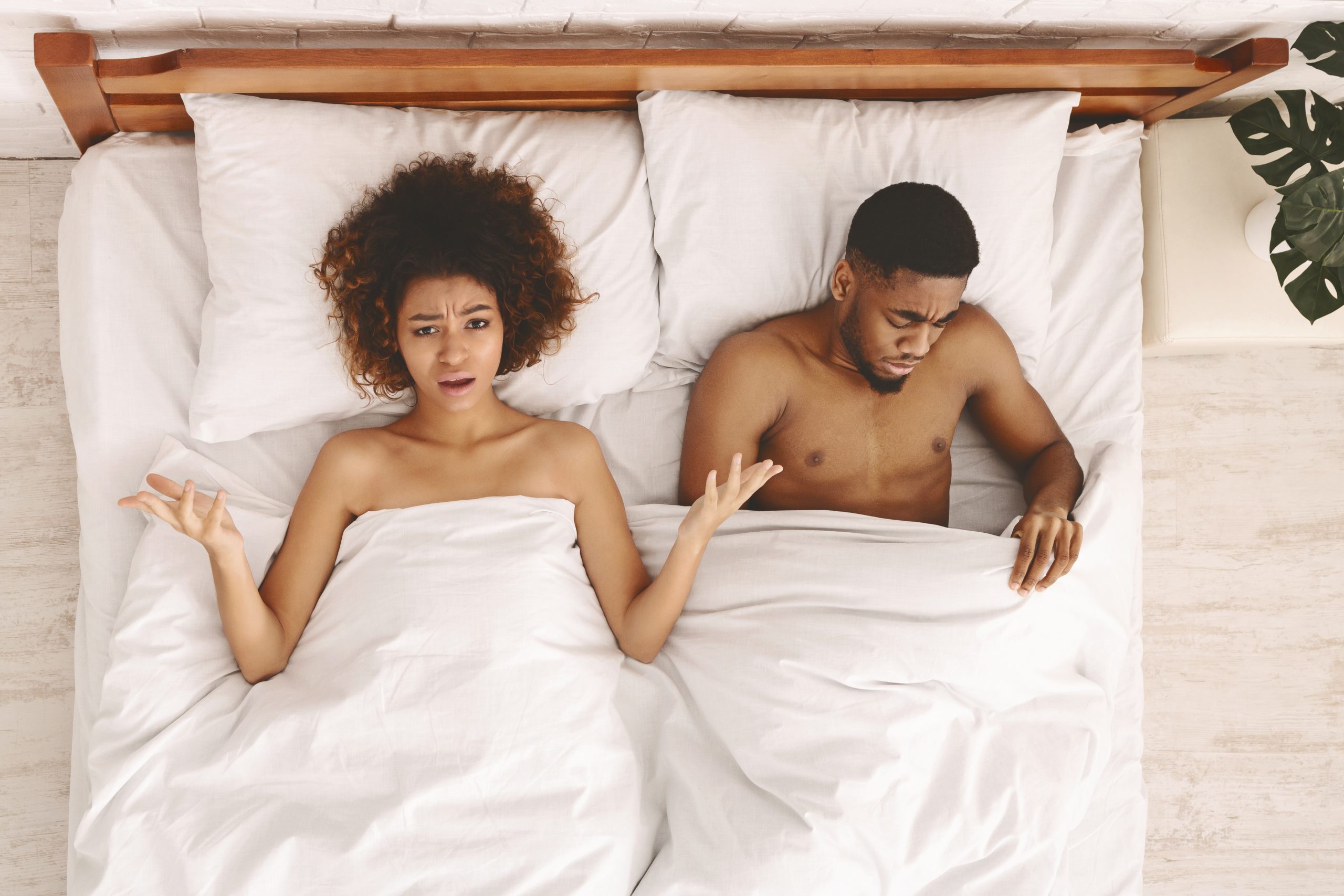 Problema sexual. Mulher negra descontente deitada na cama, homem triste a olhar por baixo do cobertor, vista de cima